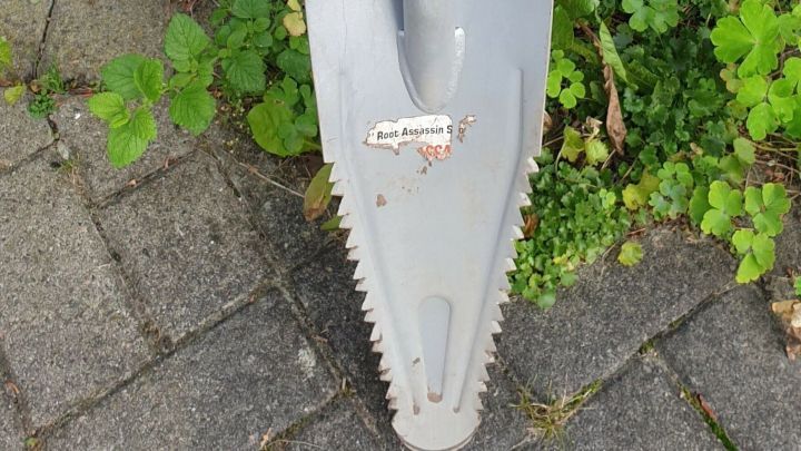 Ein spezieller Spaten mit gezackter Klinge steht auf grauem Pflaster, umgeben von gr&uuml;nen Pflanzen und einer Ziegelwand.