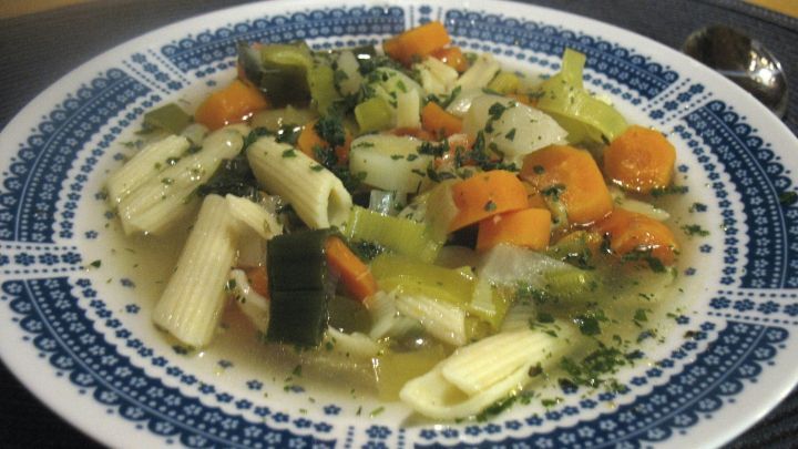 Eine bunte Karotten-Lauch-Suppe mit Gem&uuml;se und Pasta in einer dekorativen Sch&uuml;ssel, serviert auf einem dunklen Untergrund.