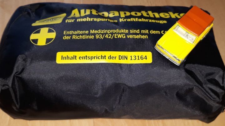 Ein schwarzer Kfz-Verbandkasten liegt auf einem Tisch, daneben ein gelbes Spielzeugauto. Der Verbandkasten zeigt wichtige Informationen.