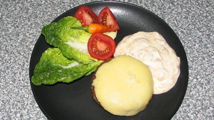 Ein gegrillter Buchweizen-Frikadelle mit geschmolzenem K&auml;se, umgeben von Salat, Tomaten und einer cremigen Sauce.