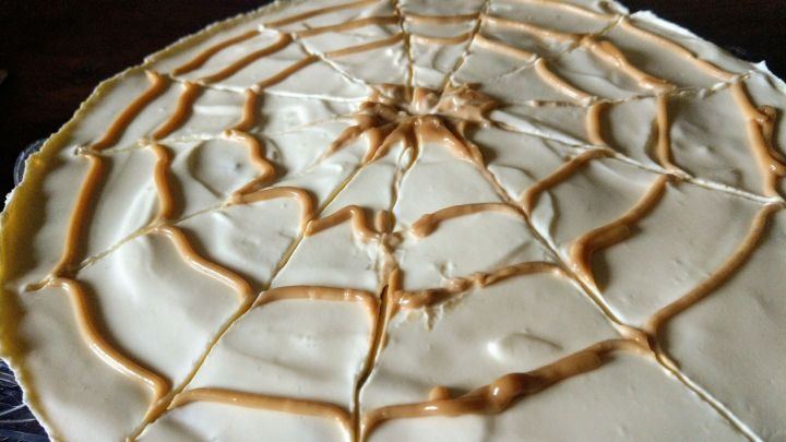 Ein runder Apfelkuchen mit einer cremefarbenen Oberfl&auml;che, dekoriert mit einem Spinnennetzmuster aus Karamell.