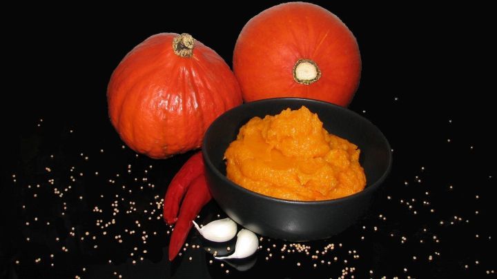 Ein schwarzes Sch&auml;lchen mit K&uuml;rbisp&uuml;ree steht vor leuchtend roten K&uuml;rbissen auf einem dunklen Hintergrund, umgeben von Gew&uuml;rzen.