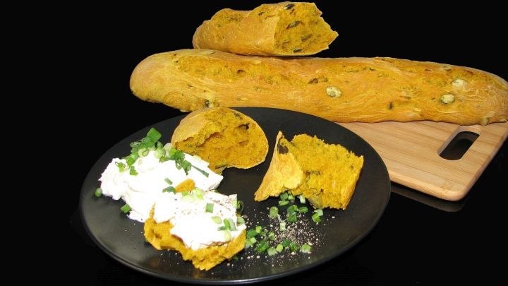 Ein goldenes K&uuml;rbisbaguette liegt hinter einem dunklen Teller, auf dem das Brot mit Kr&auml;uterquark und K&uuml;rbiskernen serviert wird.