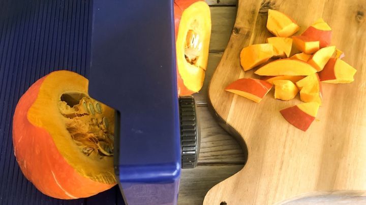 Ein orangefarbener K&uuml;rbis wird mit einem blauen Gem&uuml;seschneider in gleichm&auml;&szlig;ige St&uuml;cke geschnitten, die auf einem Holzbrett liegen.