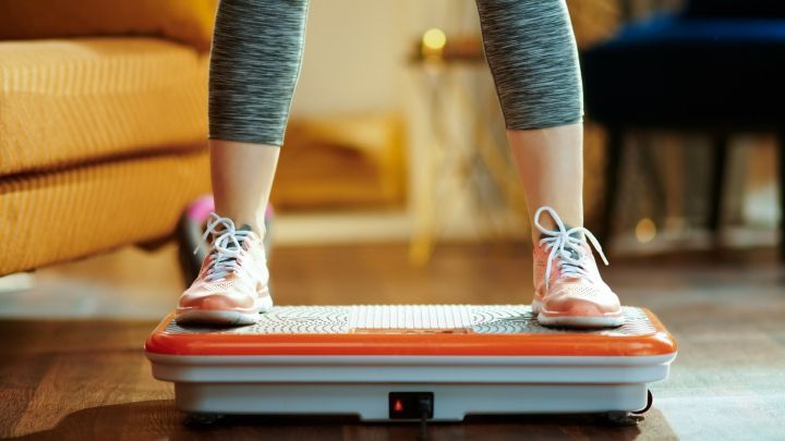 Eine Person steht auf einer Vibrationsplatte und trainiert in einem modernen Wohnbereich mit hellem Holzfu&szlig;boden.