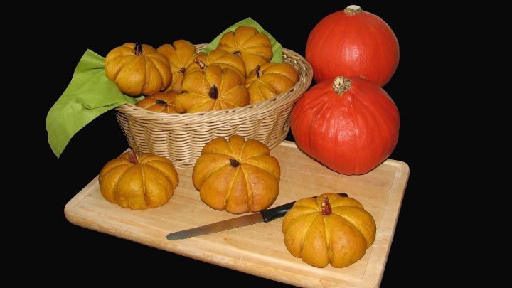 K&uuml;rbisbr&ouml;tchen liegen auf einem Holzbrett, w&auml;hrend ein Messer daneben liegt und ein Korb mit mehr Br&ouml;tchen im Hintergrund steht.