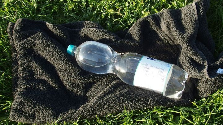Eine Wasserflasche liegt auf einem schwarzen Handtuch im gr&uuml;nen Gras, um Getr&auml;nke in der Sonne zu k&uuml;hlen.