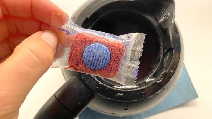 Eine Hand h&auml;lt einen Sp&uuml;lmaschinen-Tab &uuml;ber eine Kaffeekanne mit Wasser, bereit zur Reinigung.