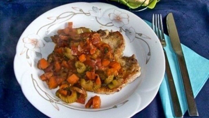 Kotelett auf einem Teller, bedeckt mit einer bunten Tomatenso&szlig;e und Gem&uuml;se, umgeben von Besteck und Br&ouml;tchen.