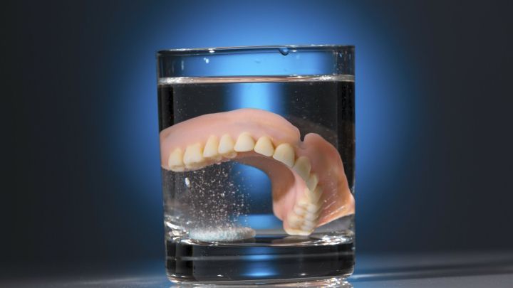 Eine Zahnprothese schwebt in einem Glas Wasser, w&auml;hrend Blasen aufsteigen und hinterleuchtet wird, was Reinigung symbolisiert.