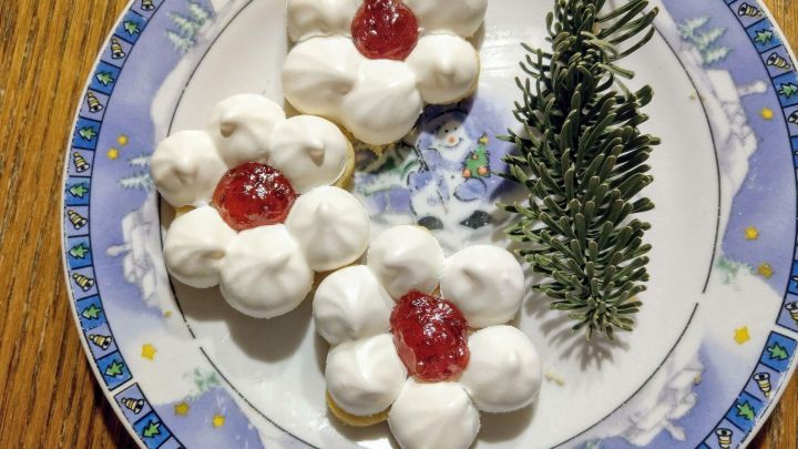 Auf einem dekorativen Teller liegen drei bl&uuml;tenf&ouml;rmige Kekse mit Sahne und Gelee, neben einem Zweig Tannengr&uuml;n.