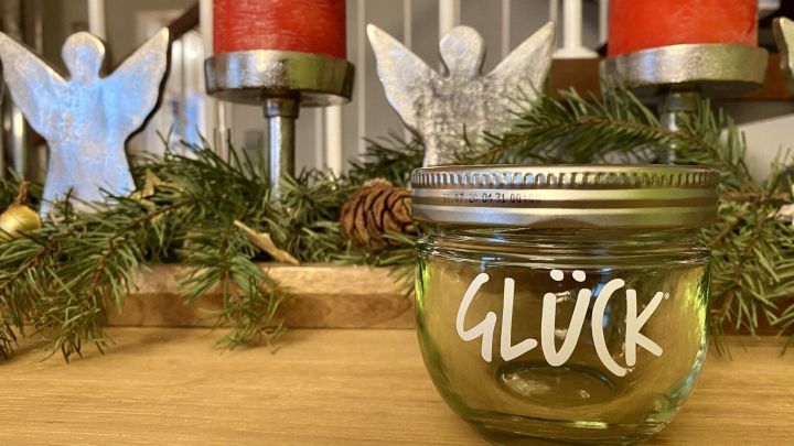 Ein beschriftetes Glas mit dem Wort &bdquo;GL&Uuml;CK&ldquo; steht auf einem Holztisch, umgeben von Weihnachtsdekor und Kerzen.