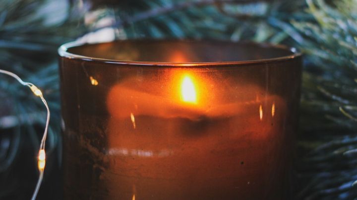 Eine brennende Kerze steht in einem rustikalen Gef&auml;&szlig;, umgeben von Tannenzweigen, was eine gem&uuml;tliche Weihnachtsstimmung vermittelt.