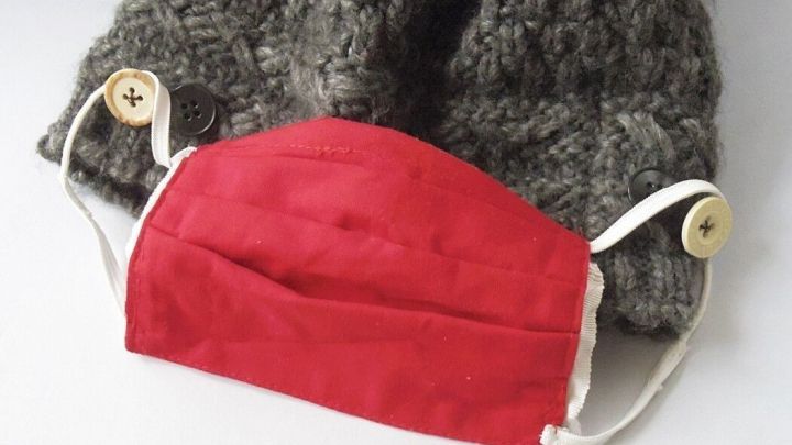 Eine rote Maske liegt auf einer gestrickten, grauen M&uuml;tze, an der Kn&ouml;pfe zum Befestigen der Maske angen&auml;ht sind.