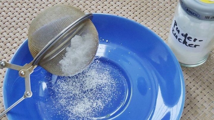 Ein feines Teesieb streut Puderzucker auf einen blauen Teller, w&auml;hrend ein Glas daneben steht.