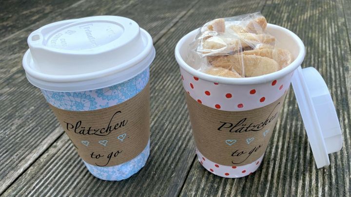 Zwei Pappbecher mit einem dekorativen Design, einer gef&uuml;llt mit Pl&auml;tzchen, stehen auf einem Holzuntergrund.