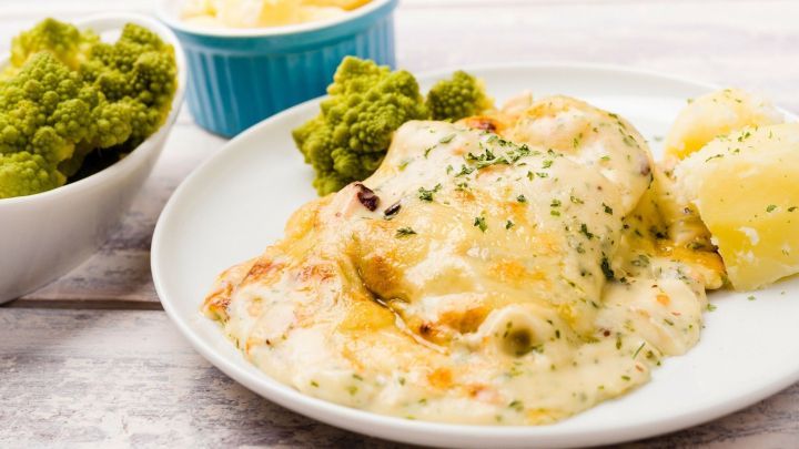 Ein goldbrauner Fischauflauf mit K&auml;sekruste liegt auf einem Teller, umgeben von Kartoffeln und gr&uuml;nem Gem&uuml;se.