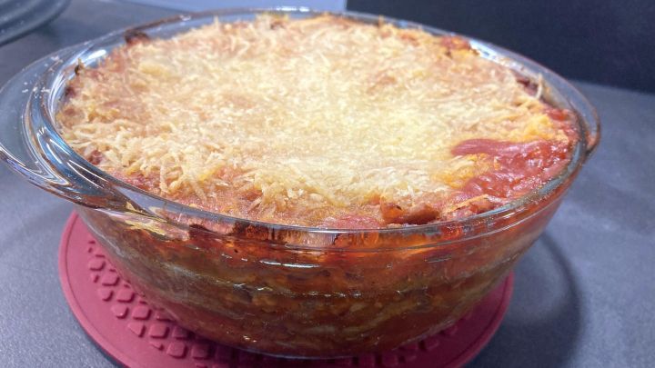 Eine goldbraune vegane Lasagne in einer Glasform steht auf einem roten Untersetzer, umgeben von dunklem K&uuml;chenuntergrund.