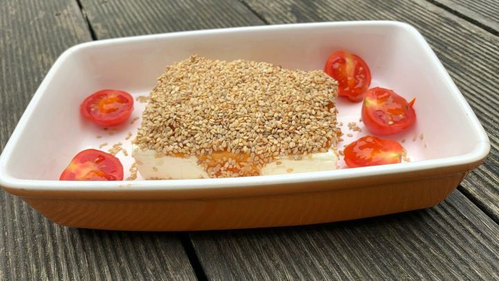 Gebackener Feta liegt in einer Schale, umgeben von Sesam und halbierten Tomaten, auf leicht verwittertem Holz.