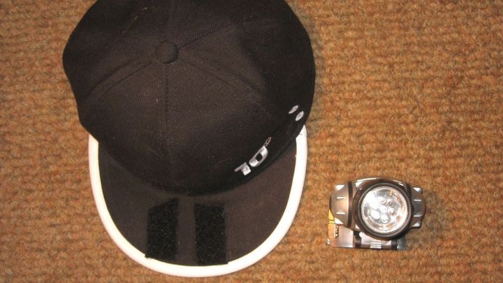 Eine schwarze Kappe liegt neben einer Stirnlampe, die zum praktischen Cap-Light umgewandelt wird, auf einem Teppichboden.