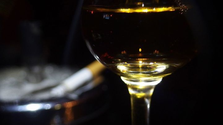 Ein Glas Wein schimmert im dunklen Raum, neben einem Aschenbecher mit brennender Zigarette, was auf eine Feier hindeutet.