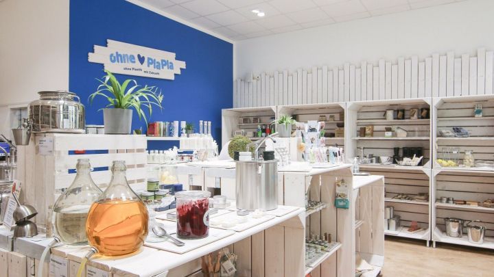 Ein Laden mit wei&szlig;en Regalen voller plastikfreier Produkte, w&auml;hrend Pflanzen und Flaschen in einem minimalistischen Stil pr&auml;sentiert werden.