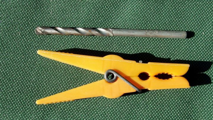 Ein gelber W&auml;scheklammer liegt neben einem Metallbohrer auf einem gr&uuml;nen Untergrund, ideal zum W&auml;sche trocknen bei Wind.