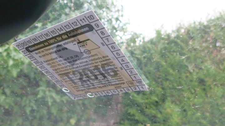 Eine Autobahn-Vignette h&auml;ngt an einer Autoscheibe, w&auml;hrend ein F&ouml;n auf sie gerichtet ist, um sie zu entfernen. Hintergrund ist unscharf.