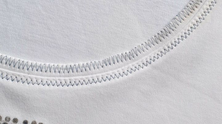 Ein wei&szlig;es T-Shirt mit einem leicht vergilbten Ausschnitt liegt flach auf einer Oberfl&auml;che, umrahmt von einem dezenten Muster.