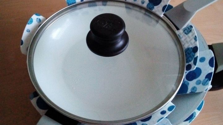 Eine Bratpfanne mit einem Deckel liegt auf einem Tisch, umgeben von farbigen Stoffschutzh&uuml;llen zum Schutz und zur Pflege.