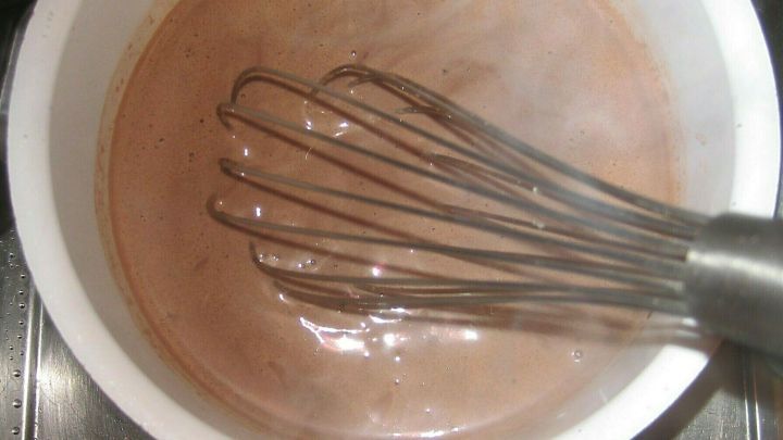 In einem wei&szlig;en Topf wird schokoladiger Pudding mit einem Schneebesen anger&uuml;hrt, w&auml;hrend Dampf aufsteigt.