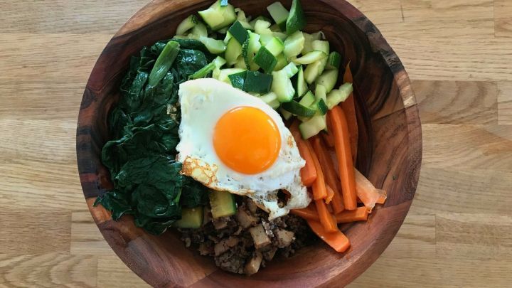 Eine Holzsch&uuml;ssel enth&auml;lt Bibimbap, mit einem Spiegelei, Gem&uuml;se und Reis, sch&ouml;n angerichtet auf einer Holzoberfl&auml;che.