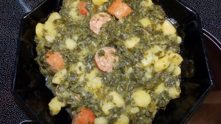 Gr&uuml;nkohl-Eintopf l&auml;uft in einer schwarzen Schale, gef&uuml;llt mit Kartoffeln und W&uuml;rstchen, auf einer Kochplatte.