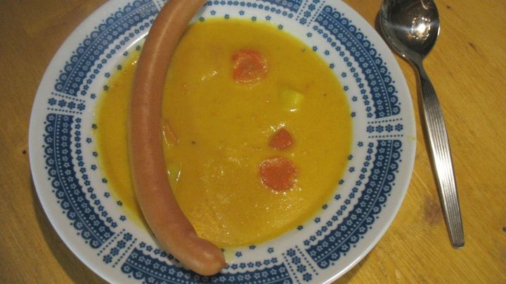 Eine dicke Karotten-Curry-Suppe mit Kokosmilch liegt in einer dekorativen Sch&uuml;ssel, neben einer Wurst und einem L&ouml;ffel.
