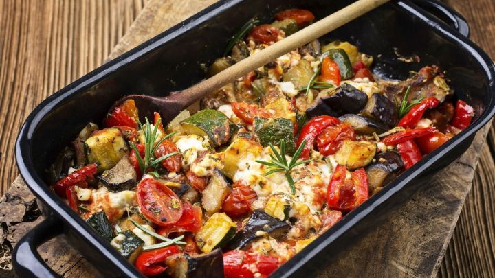 Ein bunter Gem&uuml;seauflauf wird mit einem Holzl&ouml;ffel in einer schwarzen Auflaufform pr&auml;sentiert, umgeben von rustikalem Holz.