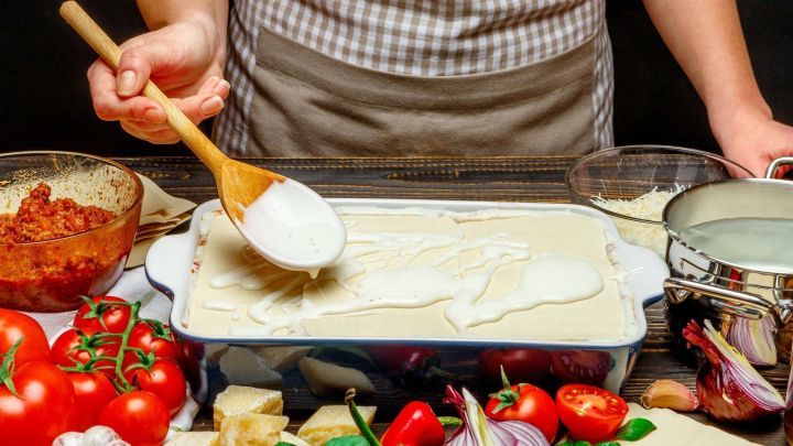 Eine Person verteilt eine cremige Lasagne-F&uuml;llung mit einem Holzl&ouml;ffel auf einer Auflaufform, umgeben von frischen Zutaten.