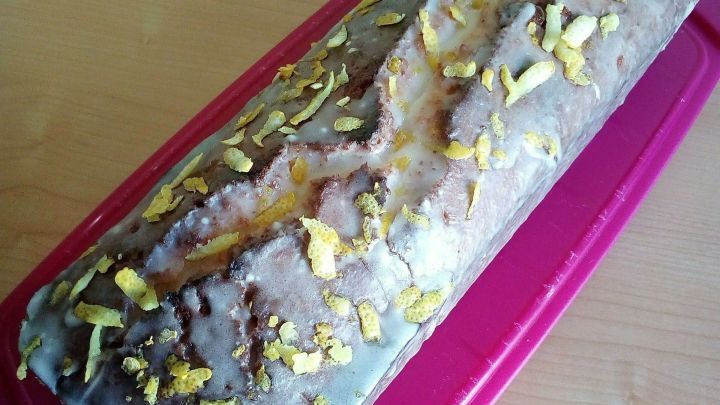 Ein Zitruskuchen liegt auf einem rosa Beh&auml;lter, glasiert und mit orangefarbener Schale dekoriert, auf einem hellen Tisch.