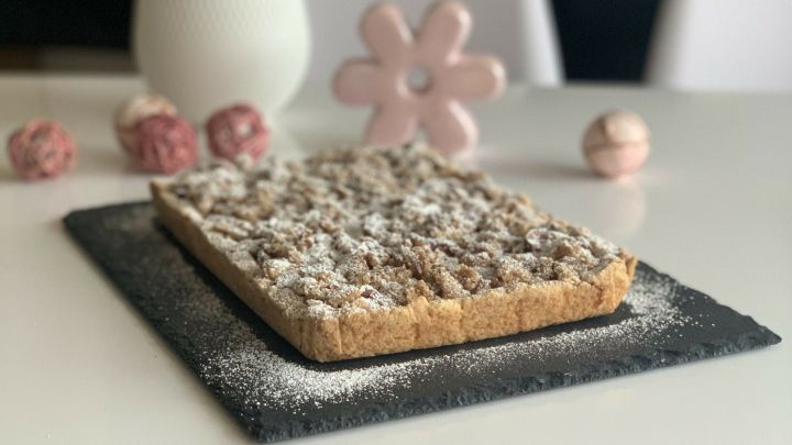 Ein Apfelstreuselkuchen mit Mandeln liegt auf einem schwarzen Teller, umgeben von einer wei&szlig;en Vase und Dekoration.