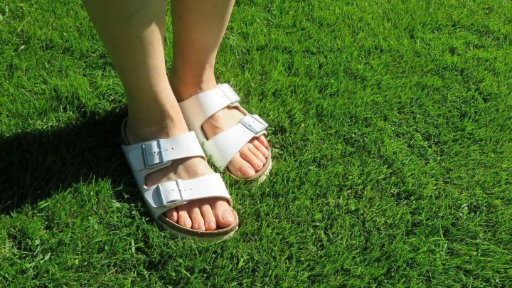 Wei&szlig;e Sandalen stehen auf gr&uuml;nem Rasen, w&auml;hrend Fu&szlig;n&auml;gel gepflegt und gesund scheinen, passend zum Thema Nagelpilz.