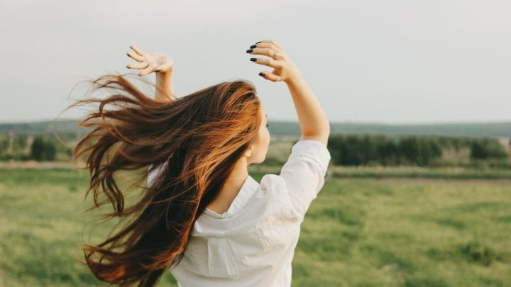 Eine Frau schwingt ihr langes, br&uuml;nettes Haar in einer gr&uuml;nen, offenen Landschaft, w&auml;hrend sie entspannend in die Ferne blickt.