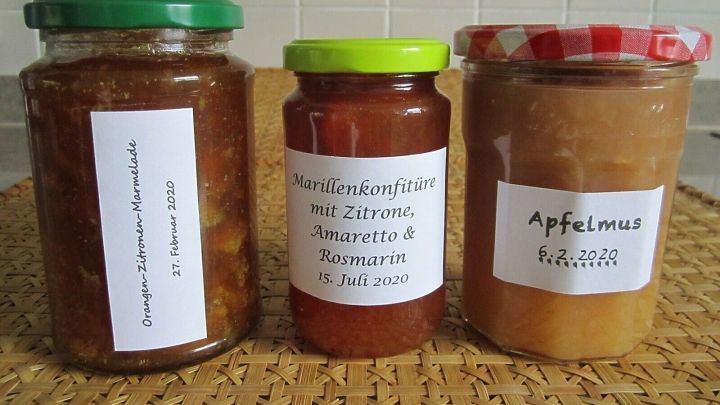 Drei beschriftete Gl&auml;ser mit Konfit&uuml;re und Marmelade stehen auf einem Tisch, umgeben von einem geflochtenen Untersetzer.