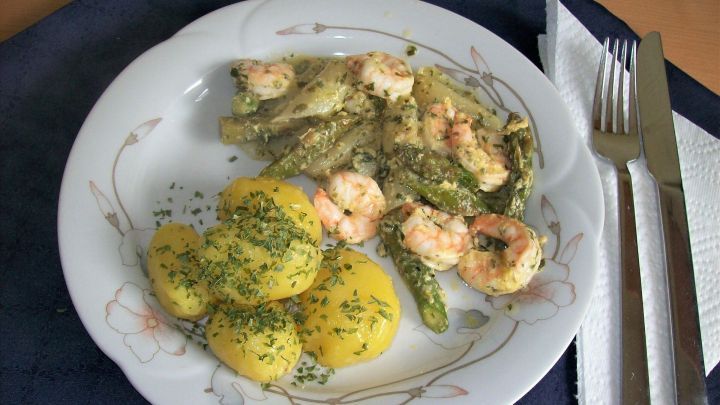 Ein Teller mit Spargel-Garnelen-Ragout liegt auf einem Tisch, umgeben von gekochten Kartoffeln und Besteck.