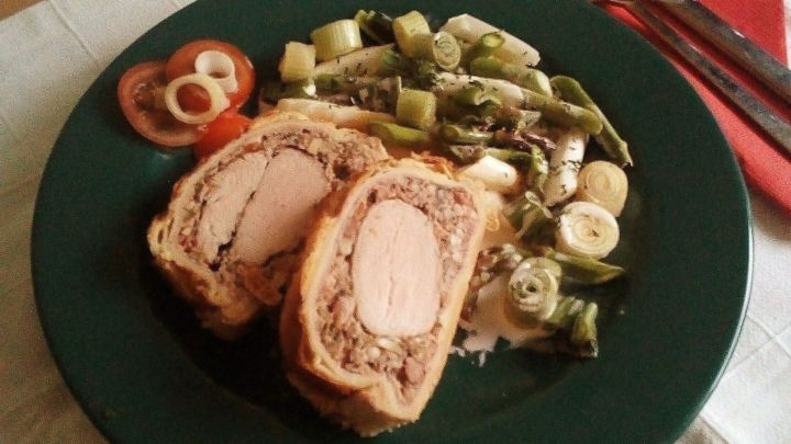 Ein saftiges Schweinefilet Wellington liegt auf einem gr&uuml;nen Teller, umgeben von frischem Spargel und Tomaten.