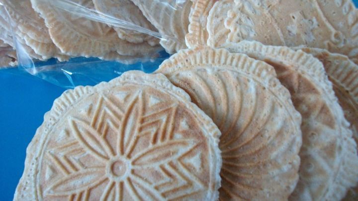 Luftige Anis-Bretzeli liegen dekorativ auf einem blauen Tisch, umgeben von weiteren h&uuml;bsch geformten Keksen in einer T&uuml;te.