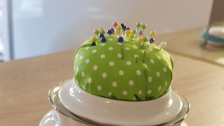 Ein Keramikgef&auml;&szlig; mit Rosenmuster h&auml;lt ein gr&uuml;nes Nadelkissen, das mit bunten Stecknadeln dekoriert ist.