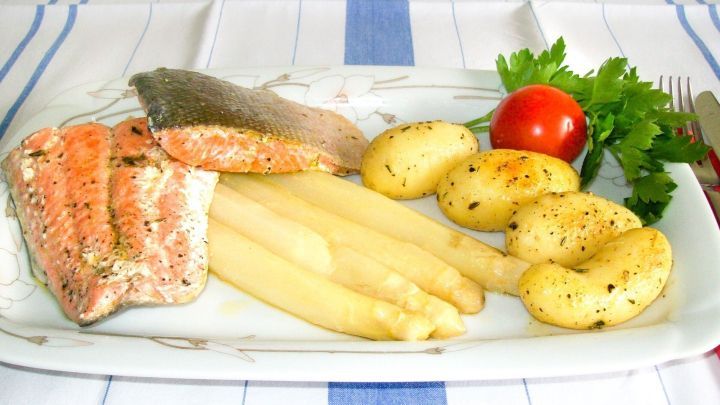 Gegrillter Wildlachs liegt auf einem Teller mit gebratenem Spargel, kleinen Kartoffeln und einem Kr&auml;uterzweig, auf Tischdecke.