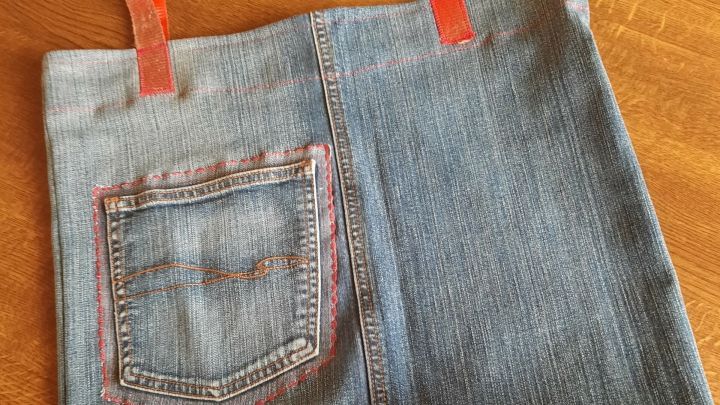 Eine handgen&auml;hte Einkaufstasche aus Jeans liegt auf einer Holzoberfl&auml;che. Die Tasche hat rot-orangefarbene Tr&auml;ger und eine aufgen&auml;hte R&uuml;ckentasche.