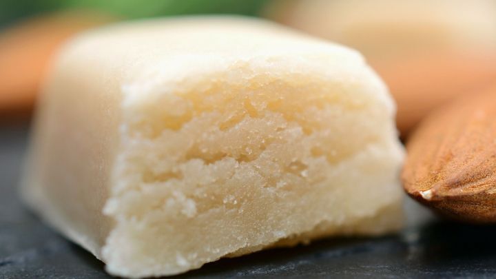 Eine St&uuml;ck Mandelmasse liegt auf einem dunklen Untergrund, umgeben von Mandeln, wobei das weiche, helle Innere sichtbar ist.