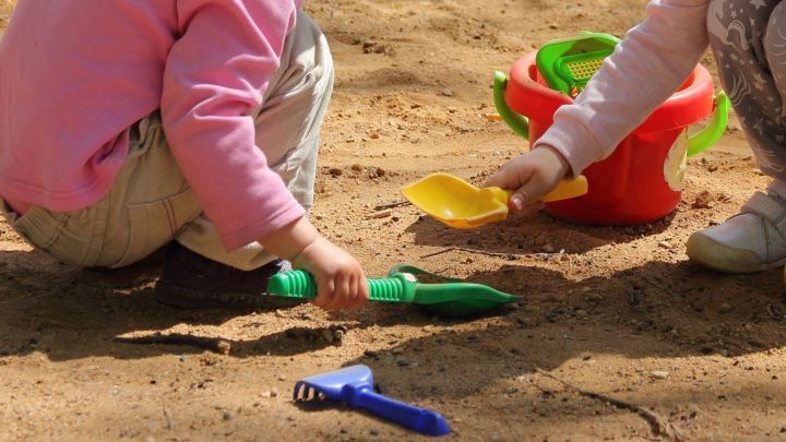 Kinder spielen im Sand, w&auml;hrend sie mit Schaufeln und einem Eimer Sandflecken auf Kleidung erzeugen.