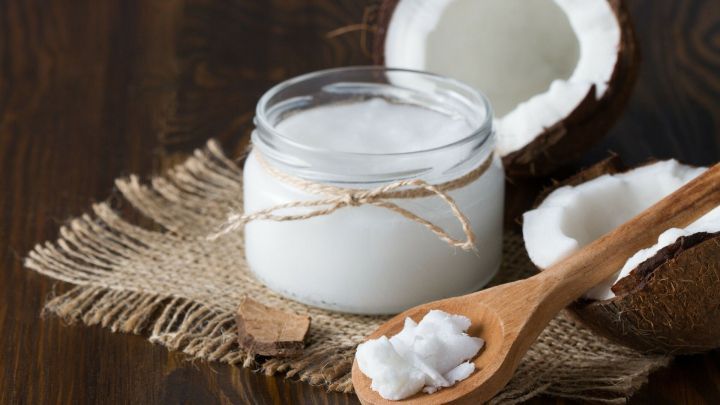 Ein Glas Kokos&ouml;l steht auf einem rustikalen Untergrund, w&auml;hrend ein Holzl&ouml;ffel mit Kokos&ouml;l daneben liegt, umgeben von Kokosn&uuml;ssen.